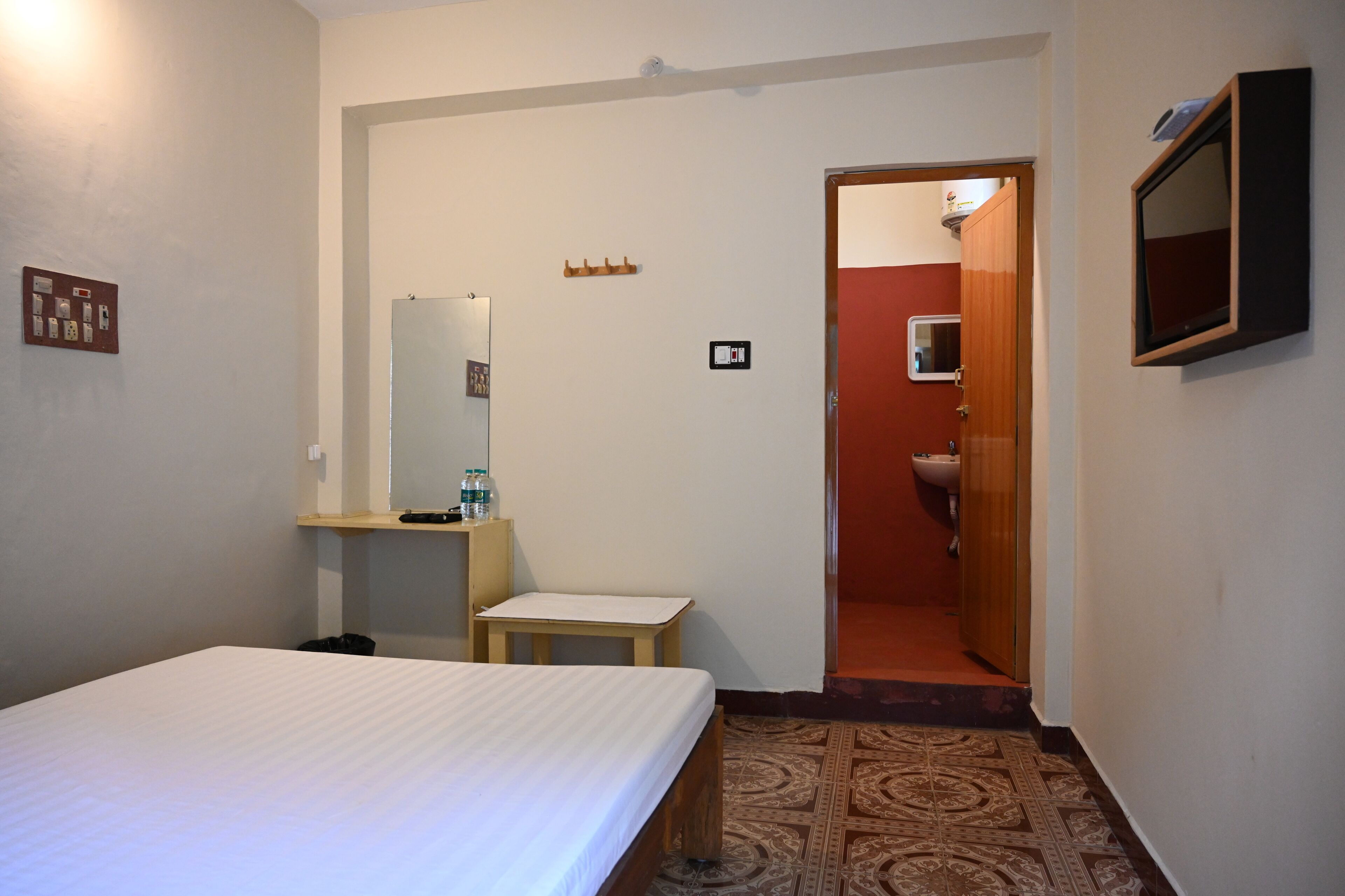 Quarto de hotel com uma cama, uma mesinha, um espelho, uma pia e uma porta que dá para outro quarto.