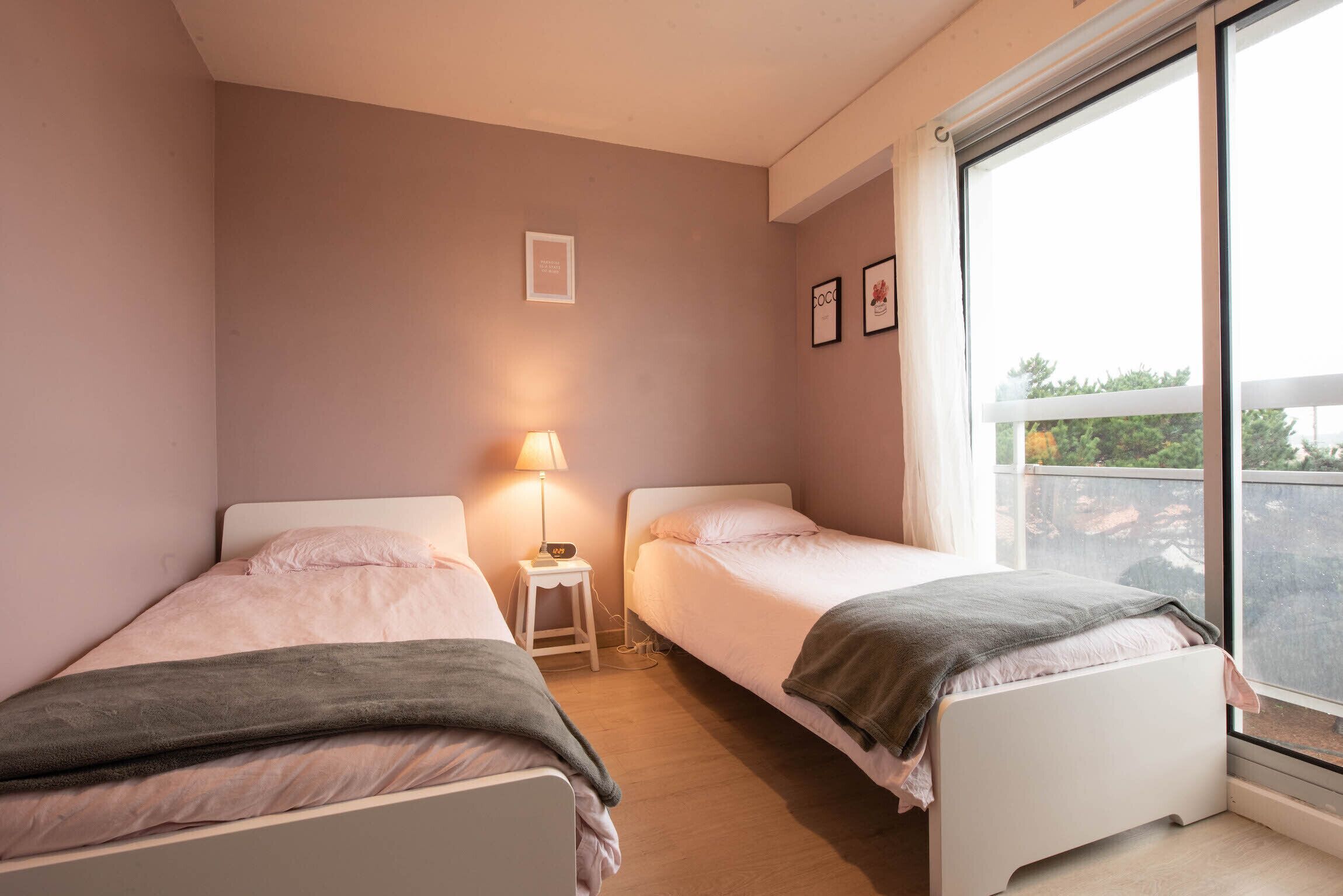 A bedroom with two beds, a nightstand with a lamp, and a window with a view of the outside.