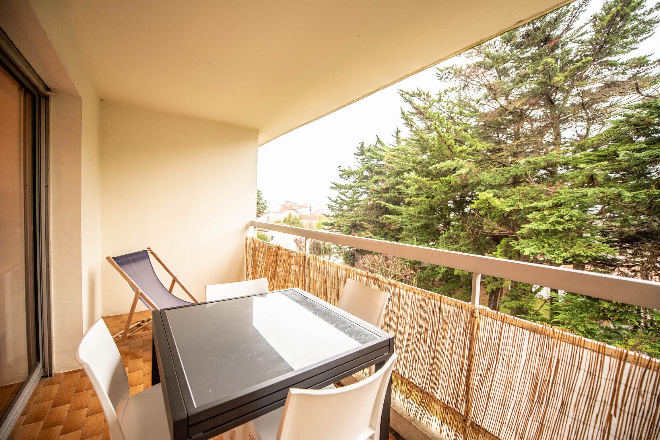 Un balcone con un tavolo e sedie in vetro, un recinto in bambù e vista sugli alberi.