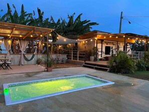 A swimming pool with a tiled deck, surrounded by outdoor seating and a covered area with string lights.