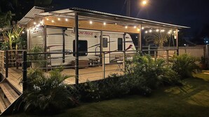 Un camping-car bien éclairé, doté d’un porche abrité et d’un espace extérieur aménagé pour s’asseoir, le tout entouré d’une végétation luxuriante.
