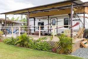 A covered outdoor area with a dining table and chairs, a hammock, and a wooden deck leading to a white RV with 'Autumn Ridge' written on it.