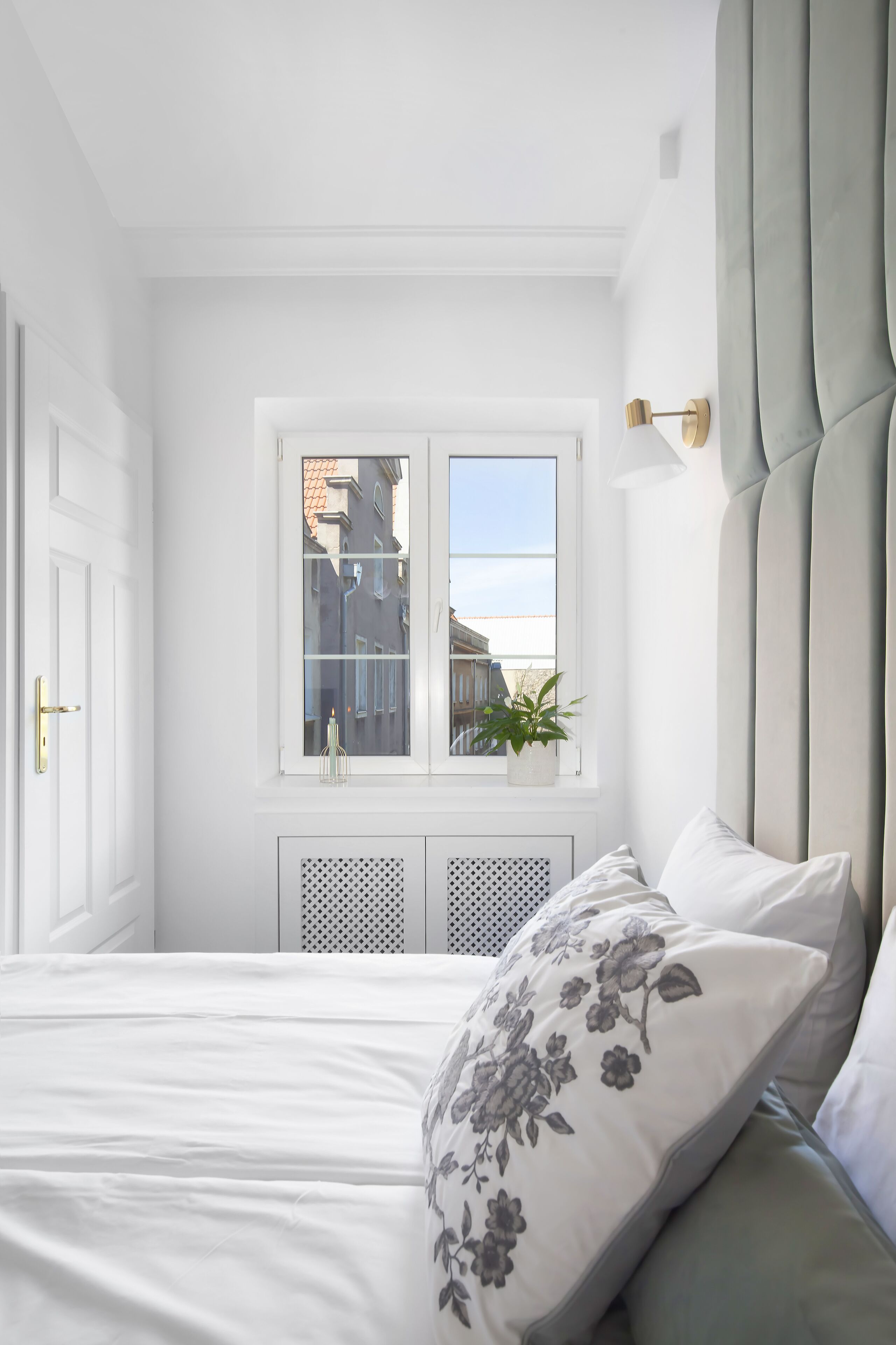 A bedroom with a bed, a window with a view of buildings, a potted plant, and a wall-mounted light fixture.