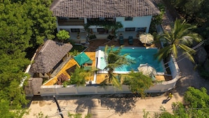 An aerial view of a resort with a swimming pool, thatched-roof structures, and lush greenery. - 6-BEDROOMS POOL VILLA in JAMBIANI (Zanzibar east coast)