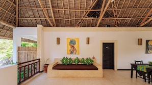 A room with a thatched roof, a sofa with a brown cushion, a dining table with a tablecloth, and a painting on the wall. - 6-BEDROOMS POOL VILLA in JAMBIANI (Zanzibar east coast)