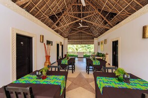 A dining area with tables set for four, each with a patterned tablecloth and a potted plant. The ceiling is thatched, and there are doors on either side of the room.