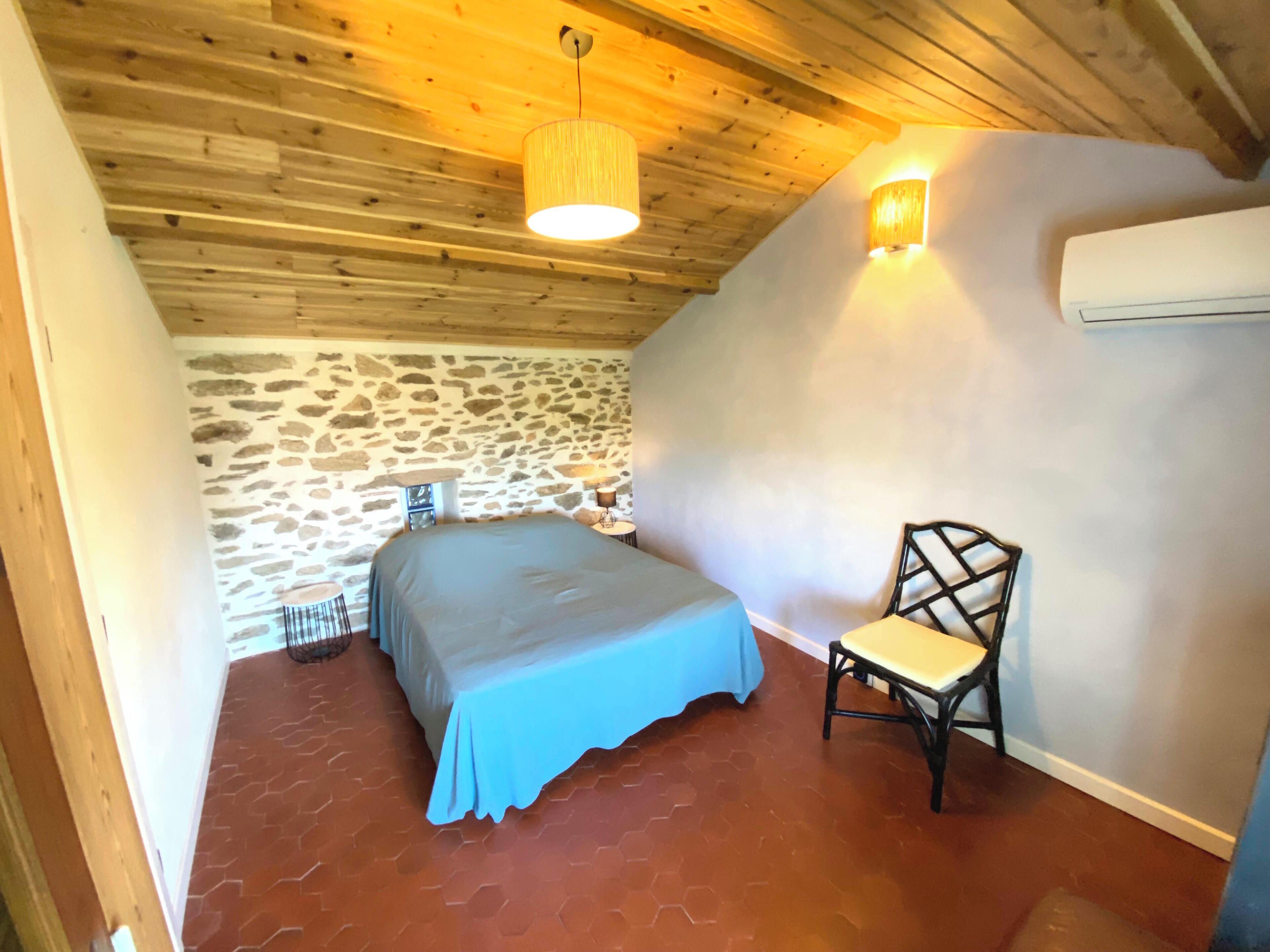 Un dormitorio con pared de piedra, una cama con cubrecama azul, una silla y techo de madera con lámpara colgante.