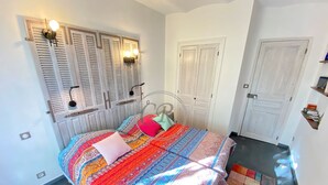 A bedroom with a bed, two windows with shutters, a door, and a small shelf with books. - LUXURY APARTMENT - T4 DUPLEX - 100 M FROM ST CLAIR BEACH (Le Lavandou)