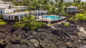 Et resort med swimmingpool omgivet af palmer, foran vulkaniske klippeformationer og fjerne bjerge.