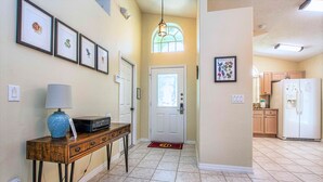 A hallway with framed pictures, a lamp, a console table, a door with a glass panel, and a kitchen area with cabinets and a refrigerator. - Cloud 9 3/4 - Southern Pool, Privacy Fence! (Kissimmee)