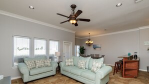A living room with two light green sofas, a ceiling fan, a chandelier, and a kitchen area with a wooden table and chairs. - Spacious Waterfront Retreat | Balcony, Huge Deck & Views (Corpus Christi)