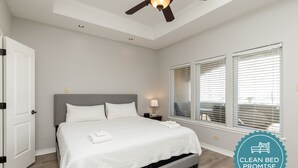 A clean, well-lit bedroom with a large bed, a ceiling fan, and a view through the window blinds. - Spacious Waterfront Retreat | Balcony, Huge Deck & Views (Corpus Christi)