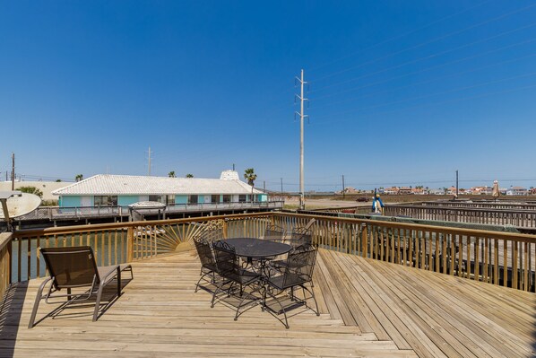 A wooden deck with a table and chairs, overlooking a beach and a building with a blue roof.