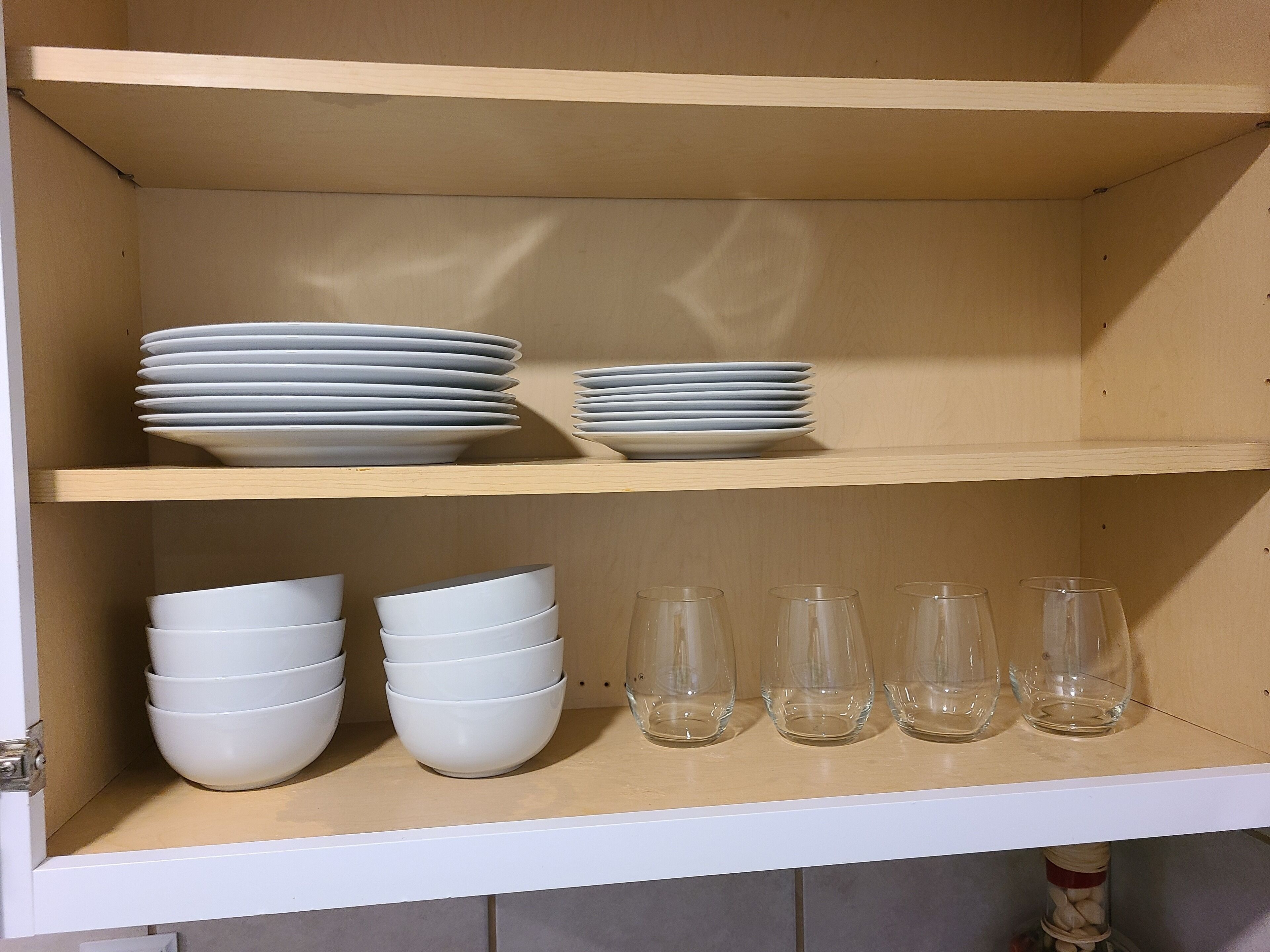 Un armario de cocina con dos estantes; el estante superior contiene platos apilados y el inferior, cuencos apilados y seis vasos transparentes.