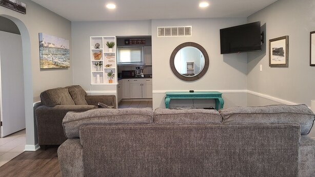 A living room with two grey sofas, a teal console table, a kitchen in the background, and a mounted TV.