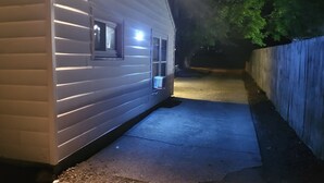 A white mobile home with a small window and a door, a concrete pathway leading to it, and a blue fence in the background.