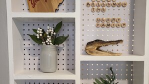 A white shelving unit with various items: a 'Youngstown' sign, a jar with white berries, a white SimpliSafe device, a potted plant, and a decorative piece spelling 'THE RAILROAD RANCH WELCOMES THE FAMILY'.