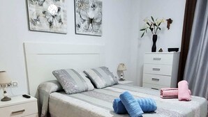 Un dormitorio ordenado con cama, almohadas, una mesita de noche, un aparador y dos cuadros en la pared.