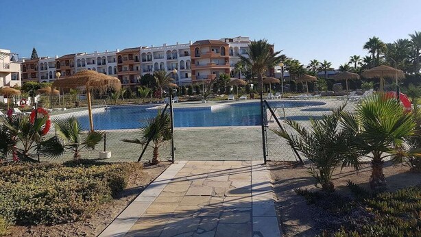 Zona de piscina en un resort, con una piscina azul cristalina, palmeras y un sendero que lleva a la piscina.