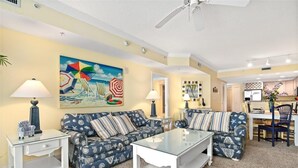 Een woonkamer met een blauwe bank met patroon, een glazen salontafel en twee bijzettafels met lampen. Aan de muur hangt een kleurrijk schilderij van strandparasols en stoelen.
