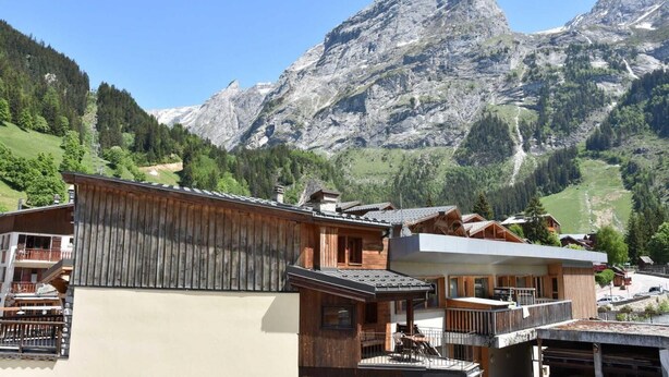 A mountain lodge with wooden architecture, a balcony with outdoor seating, and a view of a mountain range.