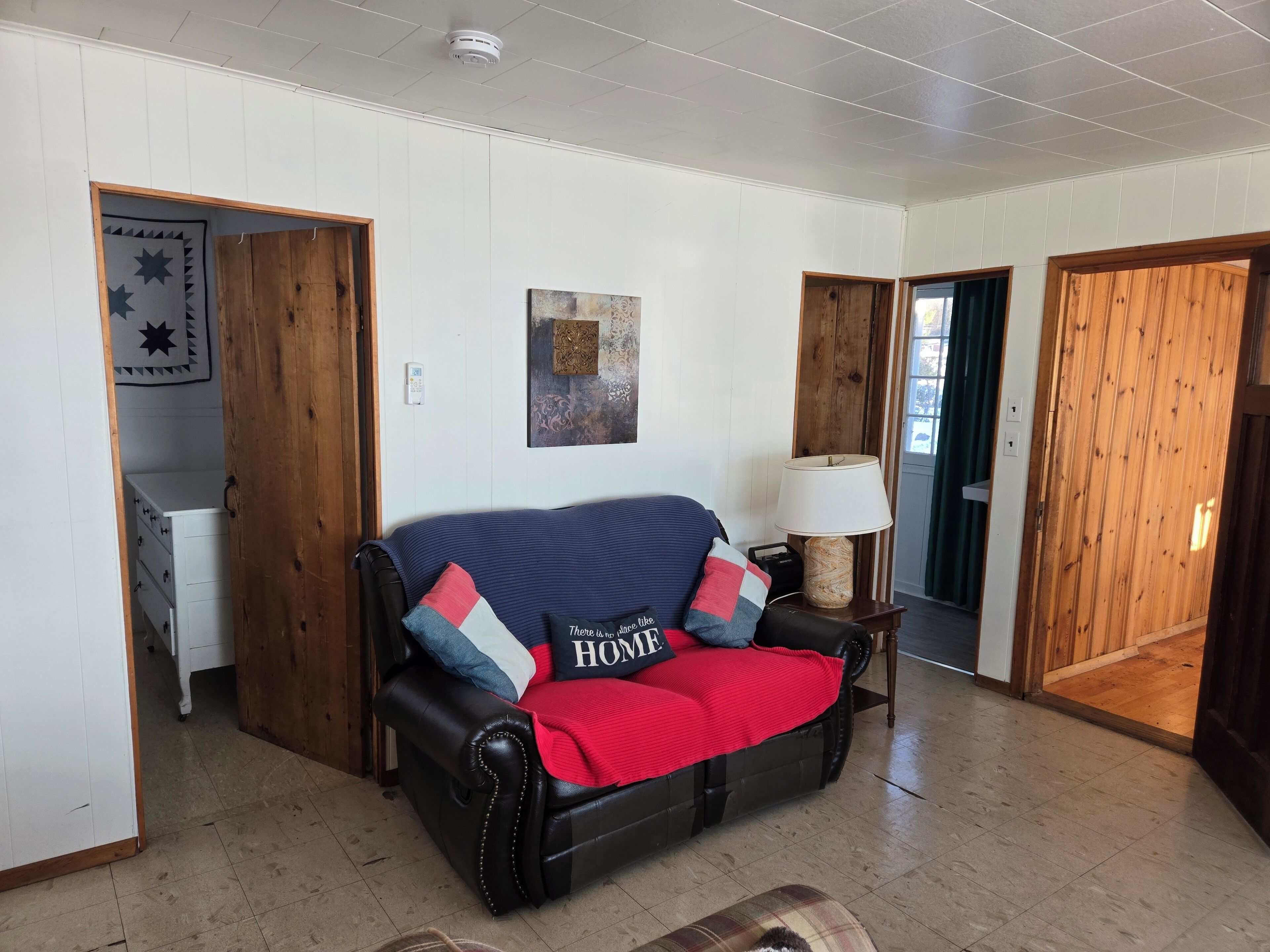 A living room with a black leather sofa, a red throw blanket, and a pillow with "HOME" text. There is a wooden cabinet and a lamp on a side table.