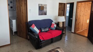 Un soggiorno con un divano in pelle nera, una coperta rossa e un cuscino con la scritta "HOME". Si nota un mobiletto in legno e una lampada su un tavolino.