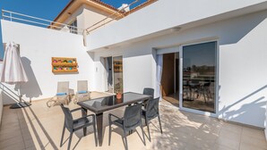 A modern outdoor patio with a glass table and chairs, a potted plant, and a view of the sea through a sliding glass door. - Narcissos 'Nissi Beach' apartment C12 (Ayia Napa)