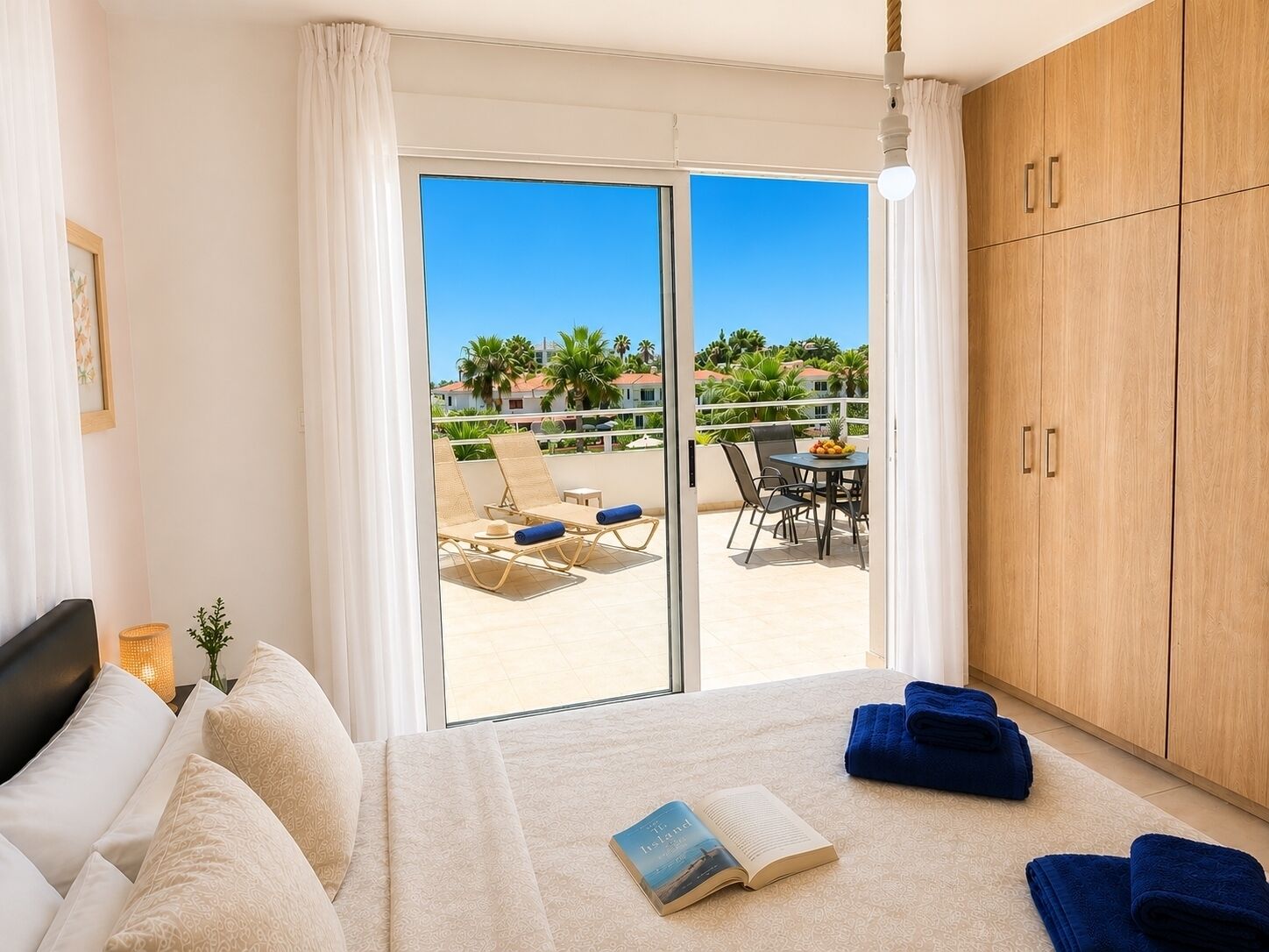 A bedroom with a bed, a reading book, a blue towel, and a view of a balcony with outdoor furniture.