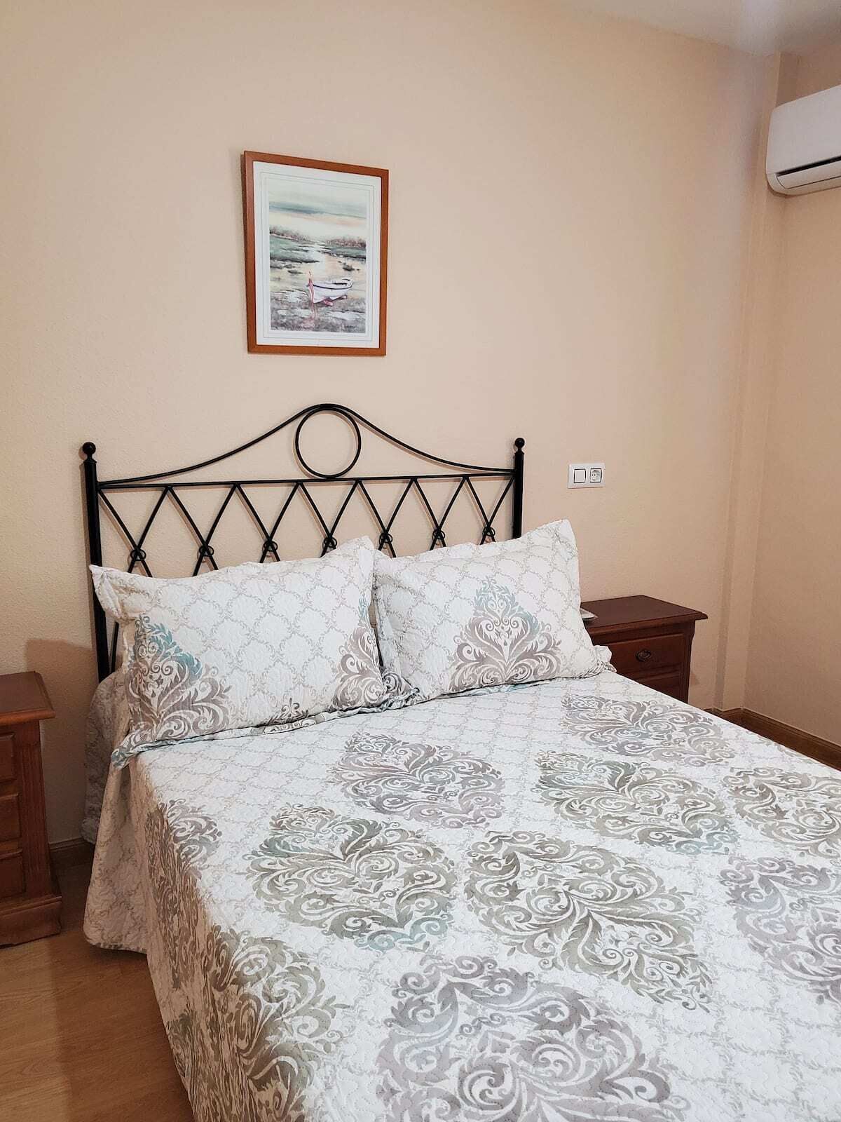 Un dormitorio con cama, mesita de noche, un cuadro enmarcado en la pared y un equipo de aire acondicionado.