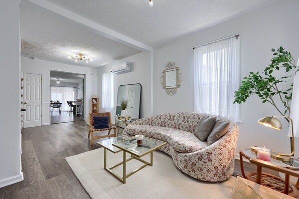 A living room with a sofa, coffee table, and a lamp. There is a dining area in the background with a table and chairs.