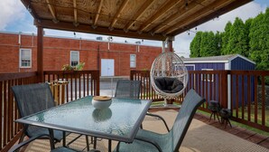 Un patio techado con una mesa y sillas de vidrio, un sillón colgante y vistas a un edificio de ladrillo al fondo.