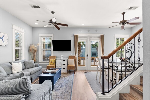 A living room with a sofa, coffee table, and a TV. There are two ceiling fans, a staircase with a railing, and a door leading to a balcony. - Steps from Pool, Mins to Beach in Blue Mountain + Free Attraction Tickets! (Santa Rosa Beach)
