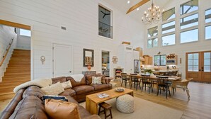 A spacious living room with a brown leather sofa, wooden coffee table, and a dining area with a wooden table and chairs. A staircase with wooden steps leads to an upper level.