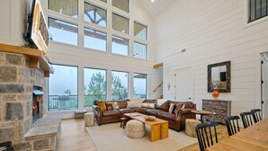 A spacious living room with a stone fireplace, a large brown leather sectional sofa, a wooden dining table, and a view of the outdoors through large windows.