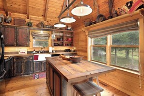 A rustic wooden cabin interior with a kitchen area, wooden cabinets, a large window, and a hanging light fixture.