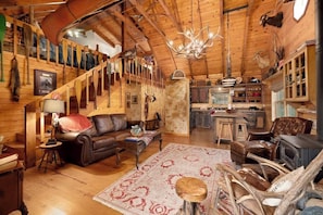 A rustic cabin interior with a wooden staircase, leather furniture, a chandelier, and a kitchen area.