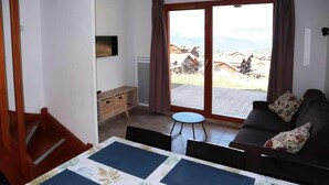 A room with a sofa, a small table, a TV, a window with curtains, and a view of a village. - Ski-in/ski-out & foot of the slopes, swimming pool, sauna, hammam, terrace, tv, ski locker, 43m² (Les Orres)