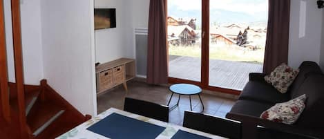 Ein Zimmer mit einer Couch, einem kleinen Tisch, einem Fernseher, einem Fenster mit Vorhängen und Blick auf ein Dorf.