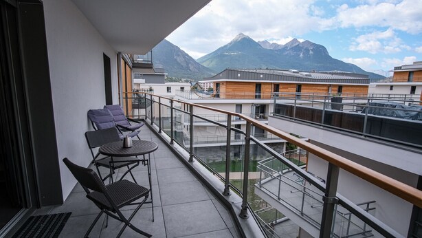 En balkon med to stole og et lille bord, udsigt over et bjerg og andre bygninger.