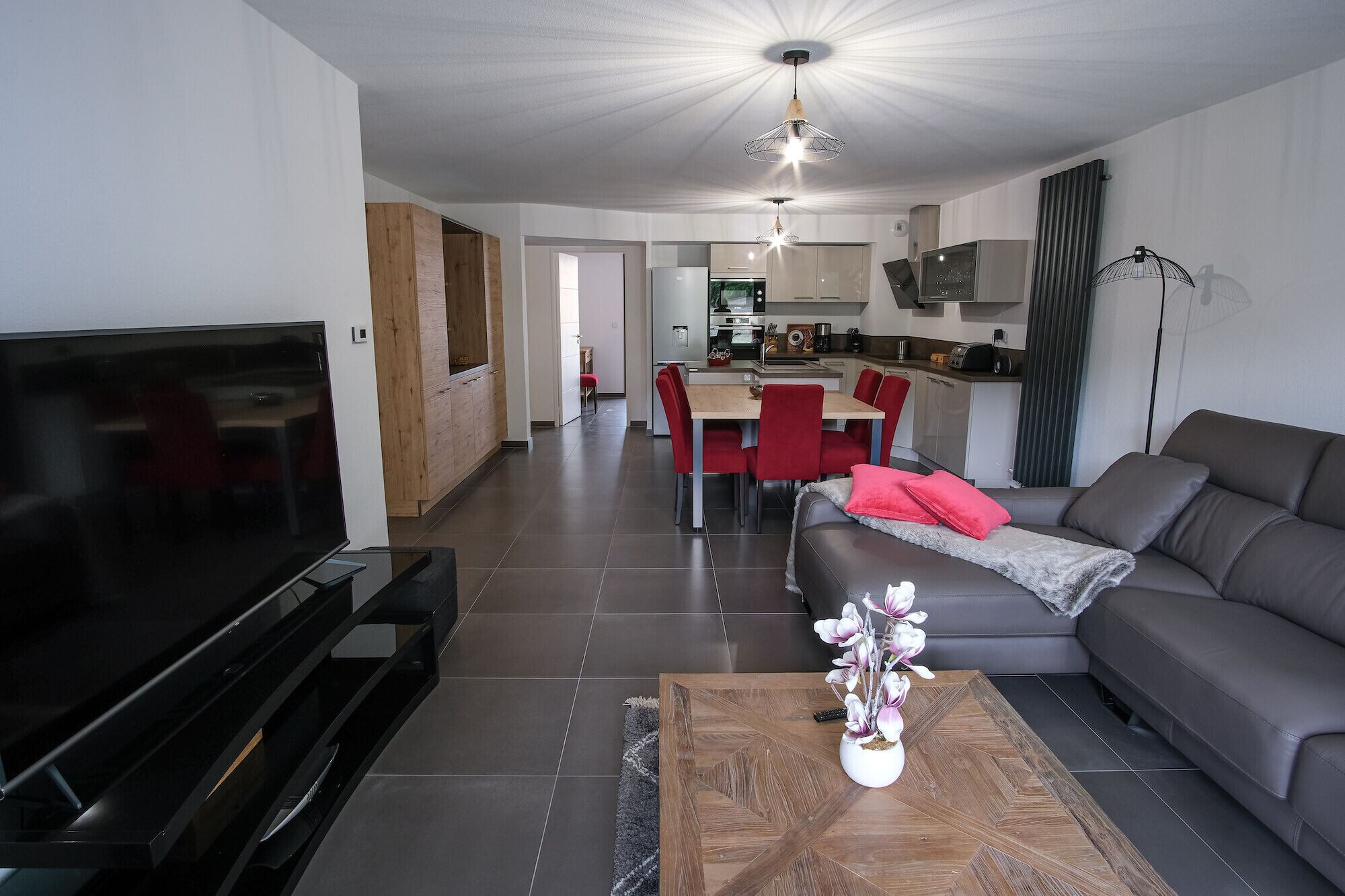 A modern living room with a flat-screen TV, a sofa, a coffee table with a plant, and a dining area with red chairs.