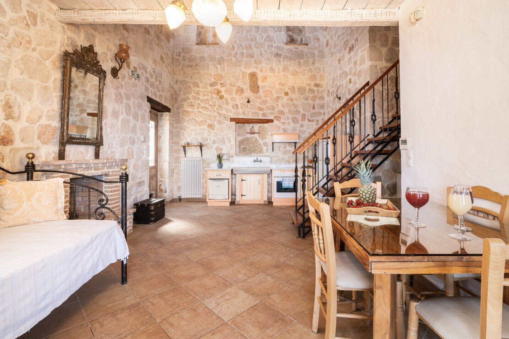 A rustic stone-walled room with a bed, a dining table with chairs, a kitchen area, and a staircase.