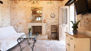 A cozy room with a stone wall, a fireplace, a white sofa, a dining table, and a TV mounted on the wall. - Strofilia Authentic Villas (Zakynthos)