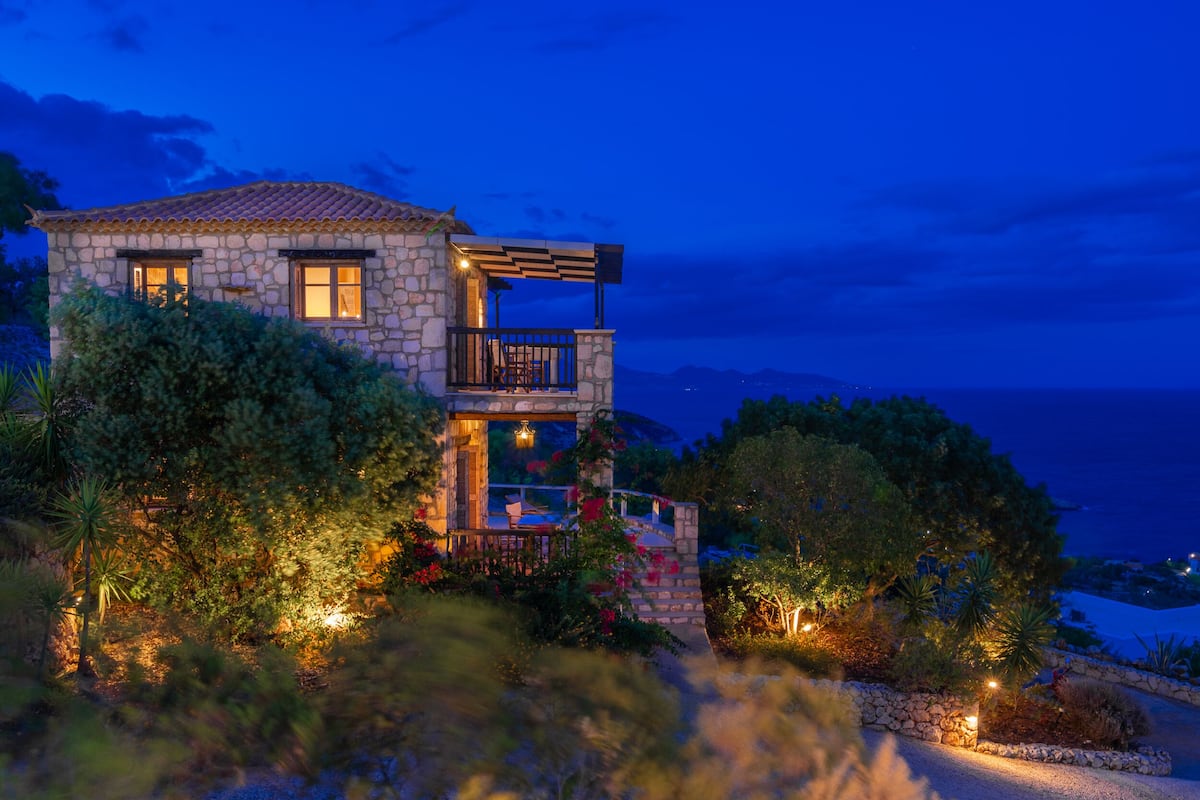 A stone house with a balcony, surrounded by lush greenery and a view of the sea.