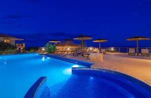 A swimming pool with lounge chairs and umbrellas, surrounded by a building with a tiled roof. - Strofilia Authentic Villas (Zakynthos)