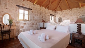 A stone-walled bedroom with a wooden ceiling, a bed with white linens, a wooden nightstand, a chair, and a mirror. - Strofilia Authentic Villas (Zakynthos)
