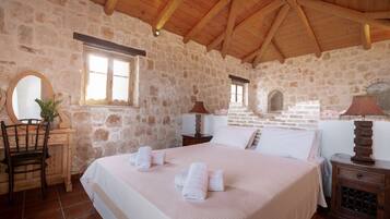 A stone-walled bedroom with a wooden ceiling, a bed with white linens, a wooden nightstand, a chair, and a mirror.