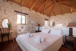 A stone-walled bedroom with a wooden ceiling, a bed with white linens, a wooden nightstand, a chair, and a mirror. - Strofilia Authentic Villas (Zakynthos)
