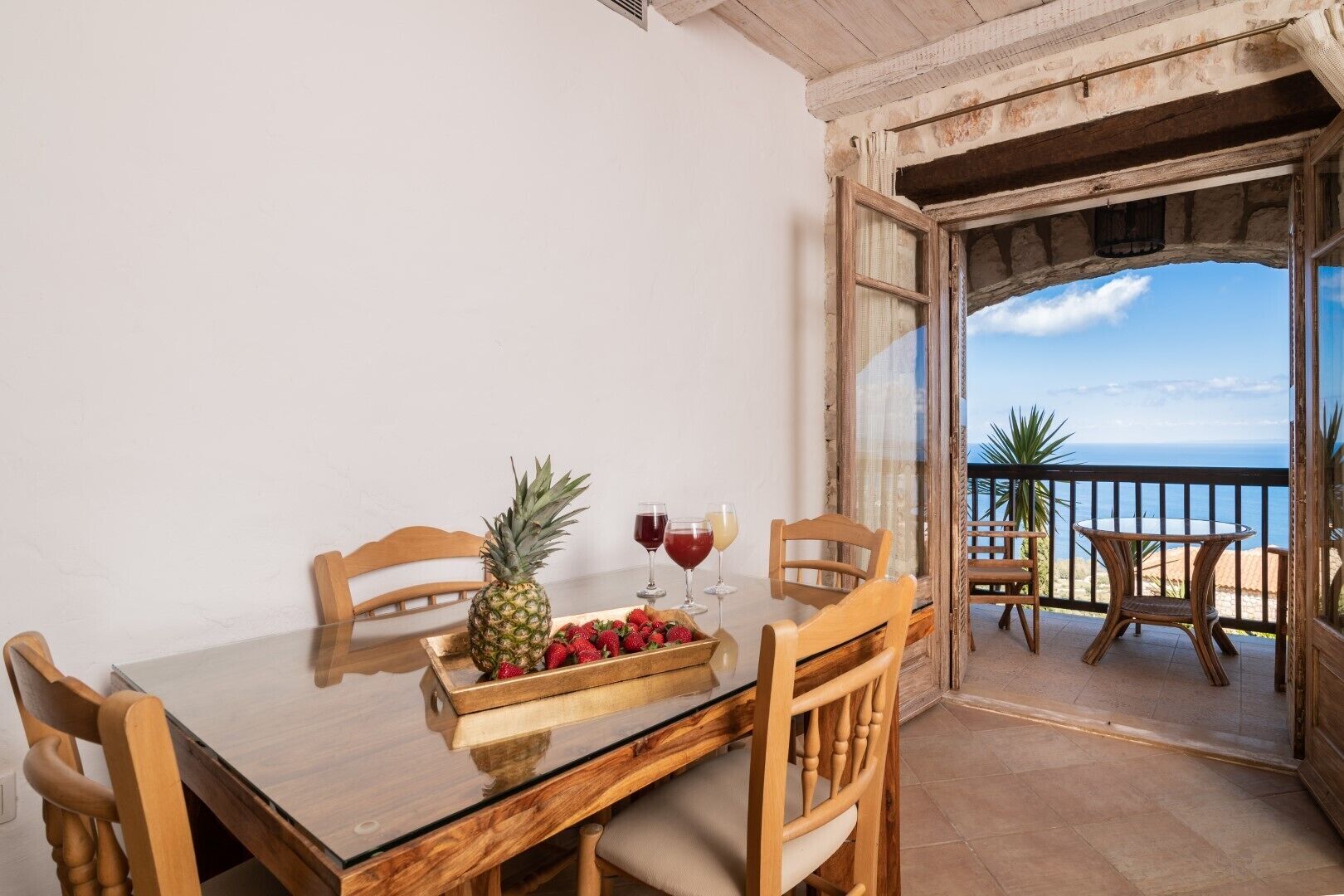 A wooden table with chairs, a bowl of fruit, and two glasses of red wine on a balcony with a view of the sea.