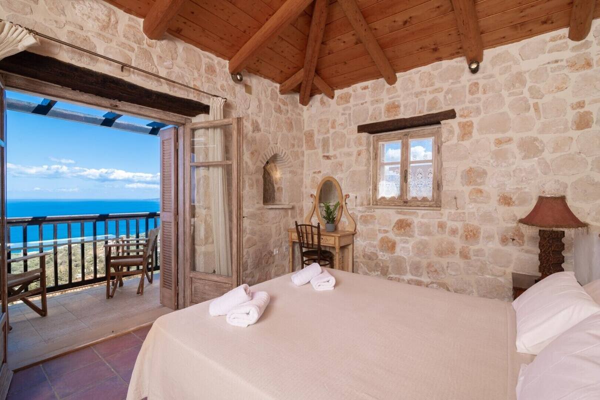A bedroom with a stone wall, a wooden ceiling, a bed with white linens, a small table with a mirror, and a view of the sea.